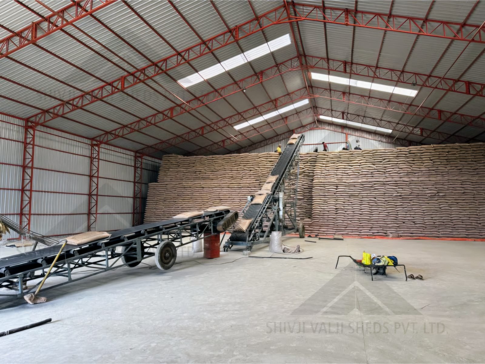Interior of a wide red-truss steel shed with white sheet roof and skylights, a tall wall of stacked grain/sugar sacks and inclined conveyor belts
