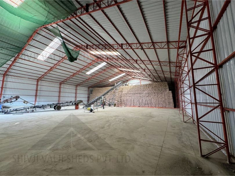 Wide interior of the storage shed from the entrance end, red lattice columns and trusses, sack stack and conveyors in the distance