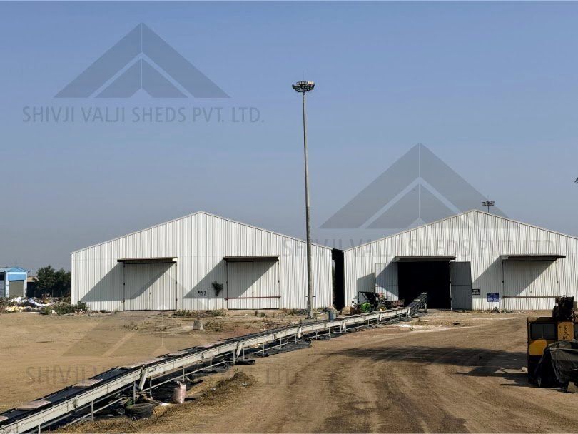 Exterior of two white colour-coated sheet gable sheds with sliding doors and canopies, conveyor and dirt road in foreground, high-mast light
