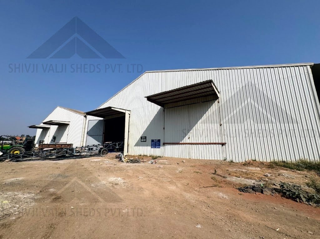 Exterior of white sheet-clad shed row with canopy doors, tractor and trusses lying at left