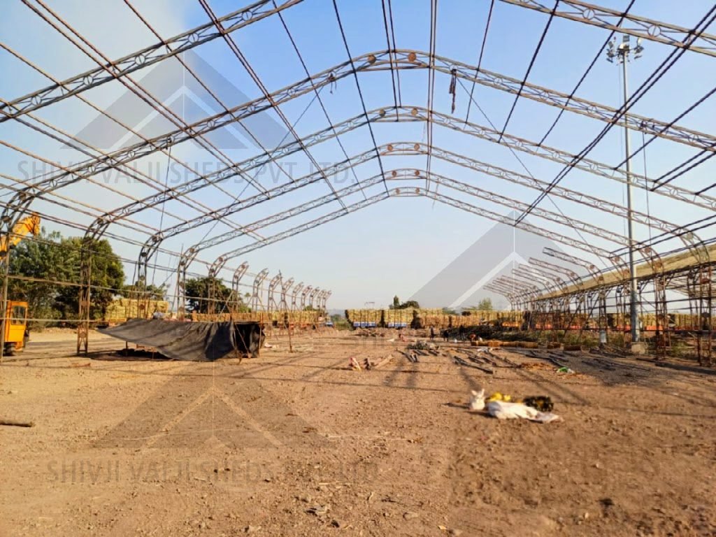 Bare arched tubular-lattice hangar frame under erection, viewed along the span, sugarcane trucks beyond (alternative: ms-steel, no cover fitted yet)