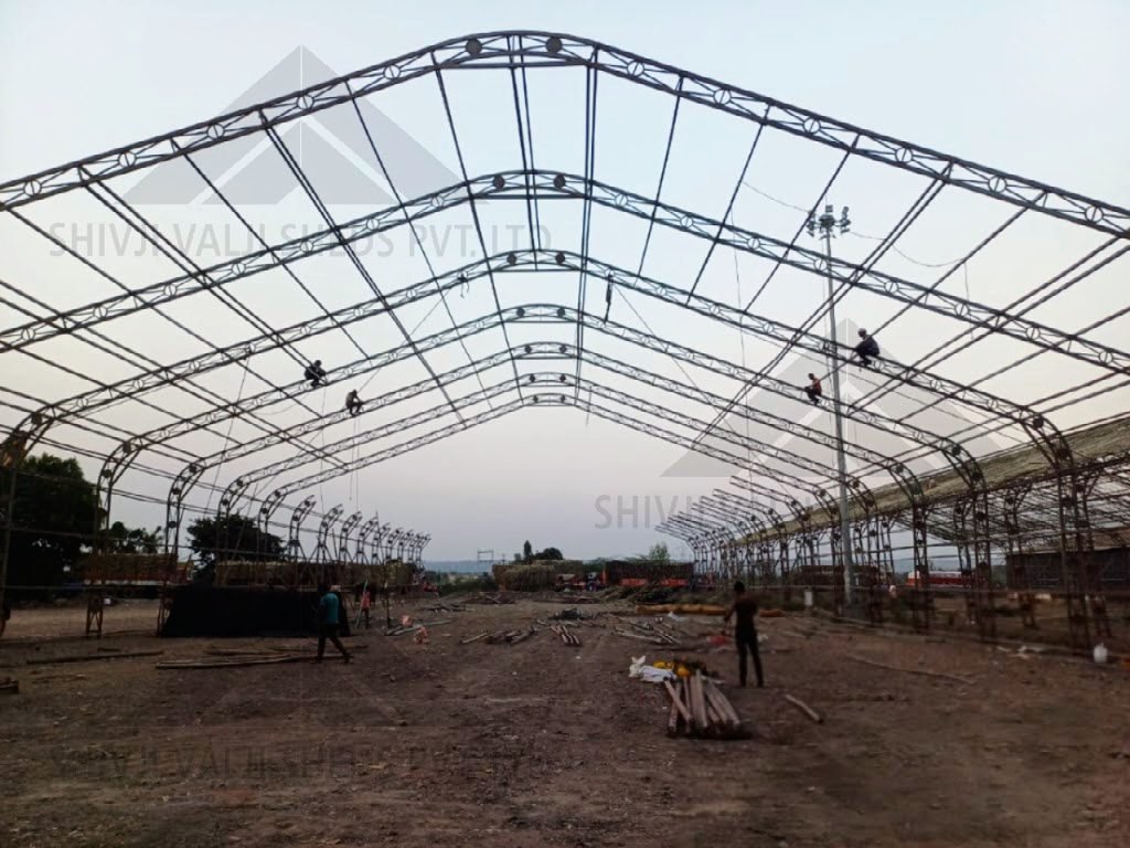 Arched lattice hangar frame under erection seen from the left side, crane at left, black tarp on ground