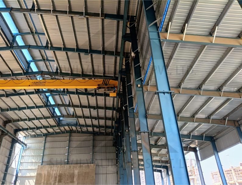 Interior of a blue-painted I-section portal-frame factory shed with a yellow EOT crane, GI roof sheet and skylights