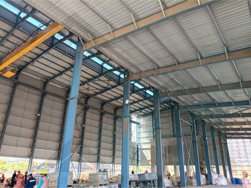 Wide interior of a large blue portal-frame factory shed with EOT crane and skylights, workers and materials on the floor