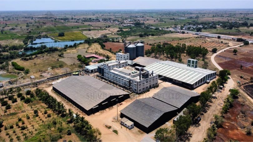 Aerial drone view of three large black tarpaulin bamboo storage sheds beside a factory with silos