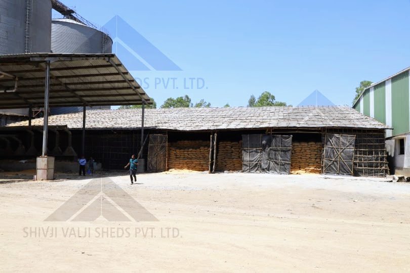 Bamboo tarpaulin storage shed full of sacks next to silos and a steel canopy, person walking in the yard