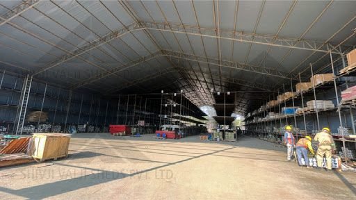 Interior of a very wide white fabric/PVC-roofed steel-truss shed with pallet racks along the sides and workers in the foreground, sunlight through the roof; low-res 512x288.