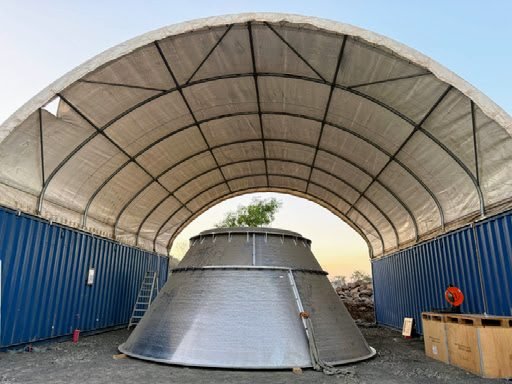 Clean frontal view of an arched white PVC dome shelter on a steel arch frame spanning two blue shipping containers, steel cone hopper underneath; low-res 512x384.