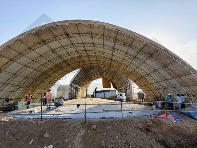 Exterior of a huge arched steel-truss hangar under construction with beige/white fabric roof, workers and trucks on the ground beneath (810x608).