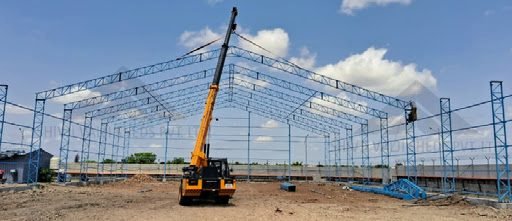 Blue-painted mild-steel lattice truss frame of a wide-span shed under erection, mobile crane lifting a truss, bare earth floor; very low-res thin crop 512x221.