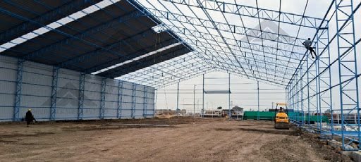 Interior of the same blue steel-truss shed with metal sheet wall at left and roof partly clad, worker on the frame and a roller compacting the floor; very low-res thin crop 512x230.
