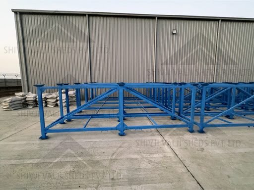 Blue-painted fabricated mild-steel frame modules laid out on a concrete yard in front of a grey metal-clad shed