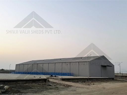 Long single-storey shed clad in grey profiled metal sheeting with a plain pitched gable roof, on a levelled open yard
