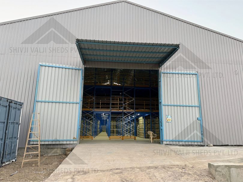 Gable end of a grey profiled-sheet shed with tall blue-framed doors swung open onto racked storage inside