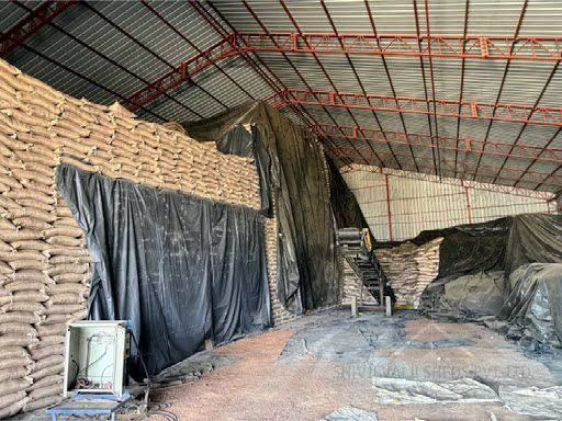 Interior of a red-primed steel truss roof shed with black tarpaulin side curtains, sacks stacked high and a conveyor